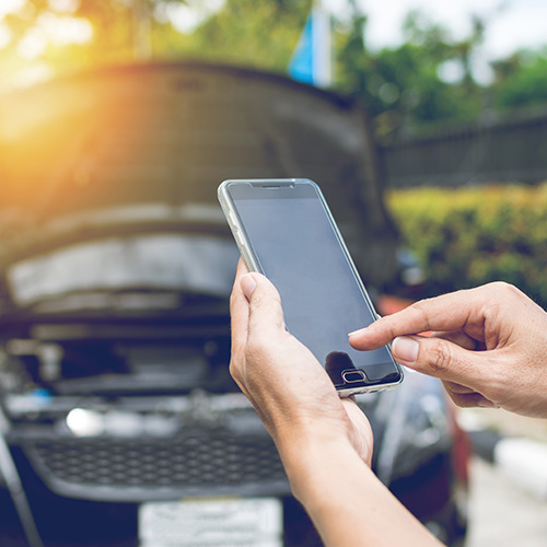 Person dialing for roadside assistance on mobile phone, car with hood lifted in the background