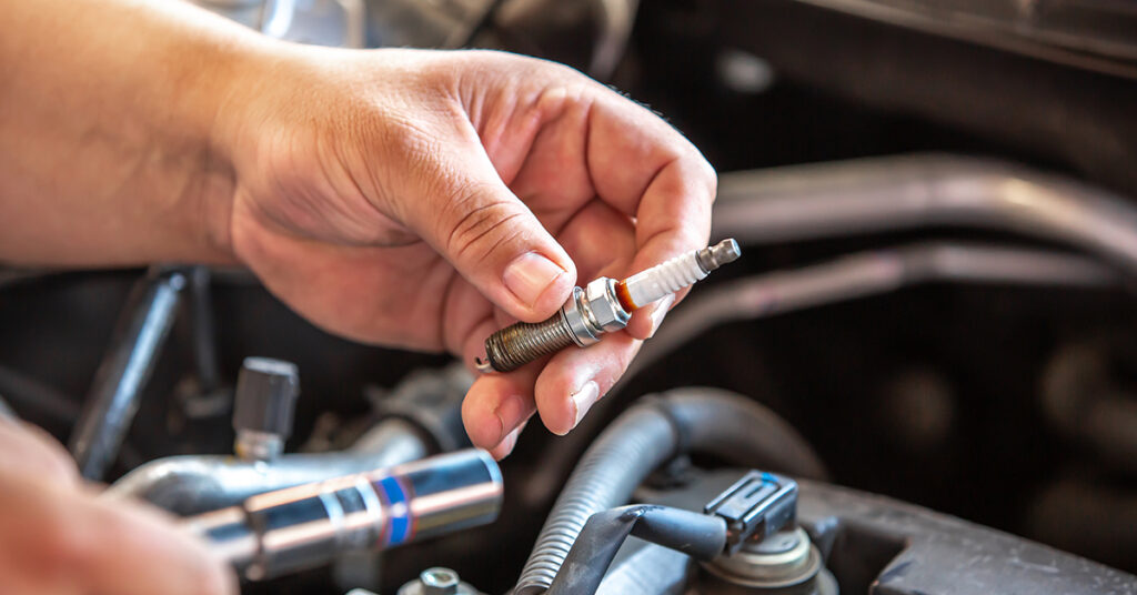 Close-up of a left hand holding a spark plug. The car engine is in the background.