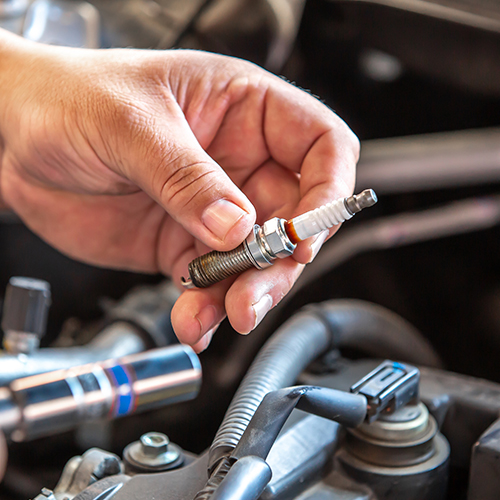 Close-up of a left hand holding a spark plug. The car engine is in the background.